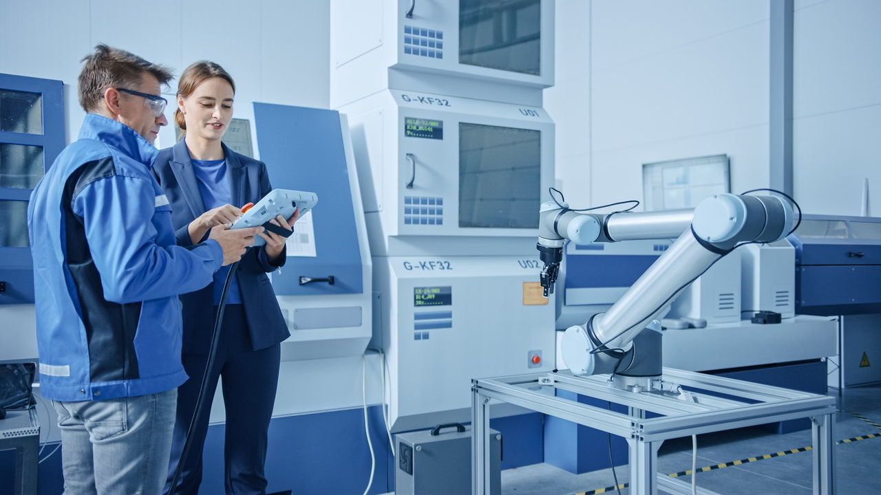 Modern Factory: Female Project Manager and Male Engineer Standing, Talking and Using Tablet Computer for Programming Robot Arm Assembly Line. Contemporary Facility with High Tech CNC Machinery