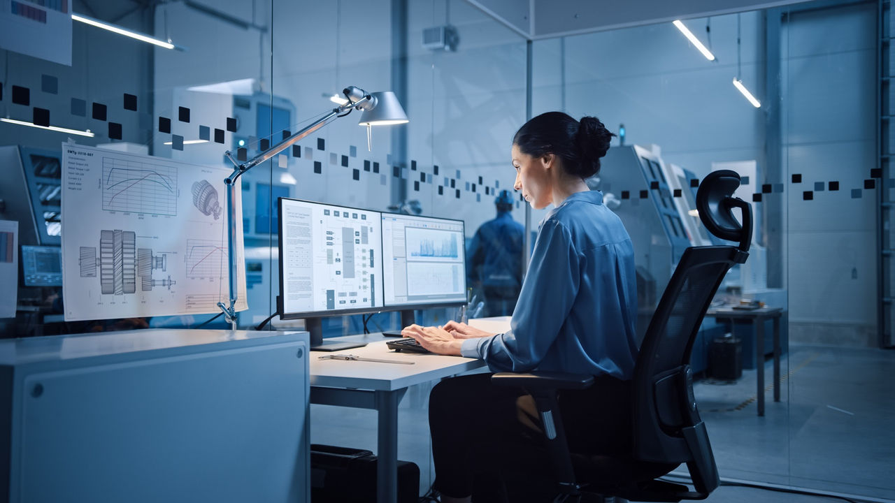 Factory Office: Portrait of Beautiful and Confident Female Industrial Engineer Working on Computer, on Screen Industrial Electronics Design Software. High Tech Facility with CNC Machinery