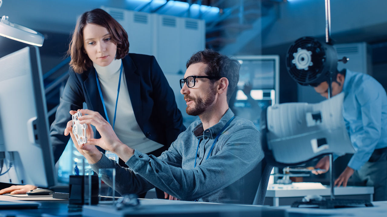 Futuristic Machine Engine Development Engineer Working on Computer at His Desk, Talks with Female Project Manager. Team of Professionals Working in the Modern Industrial Design Facility