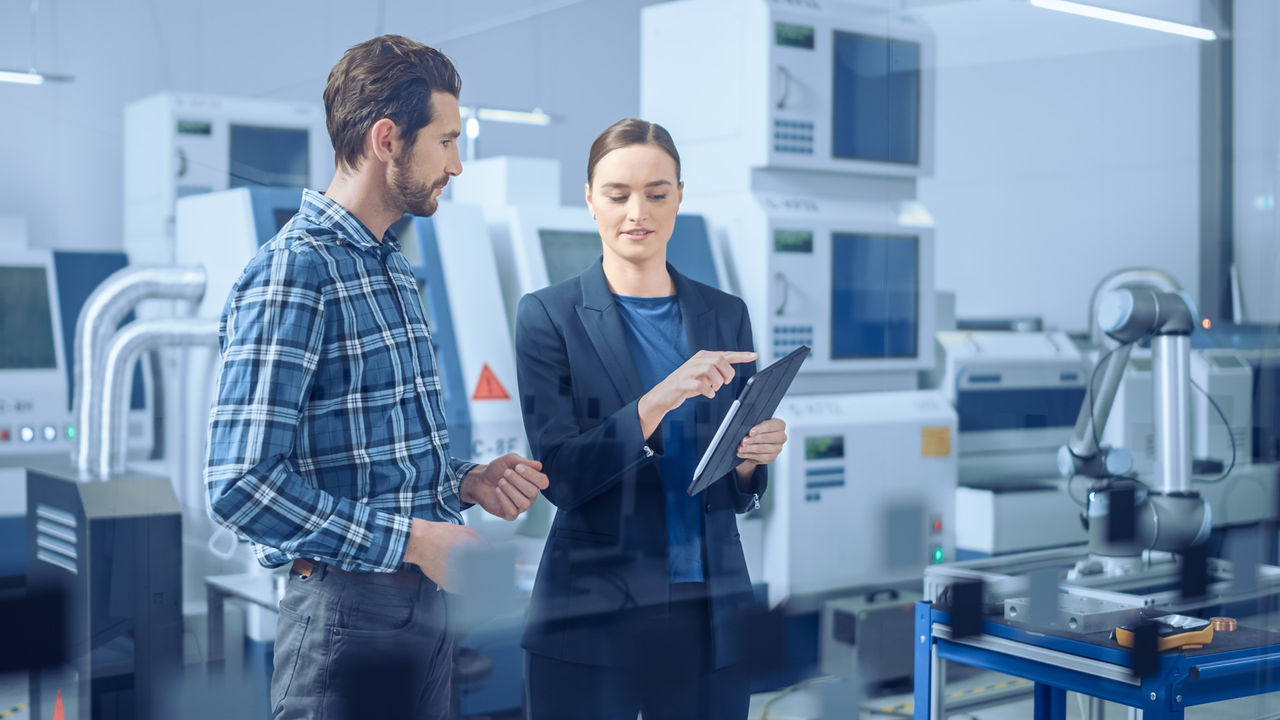 Modern Factory: Female Project Manager and Male Engineer Standing, Talking, Using Digital Tablet for Programming and Monitoring Assembly Line. Industry 4.0 Facility with High-Tech CNC Machinery